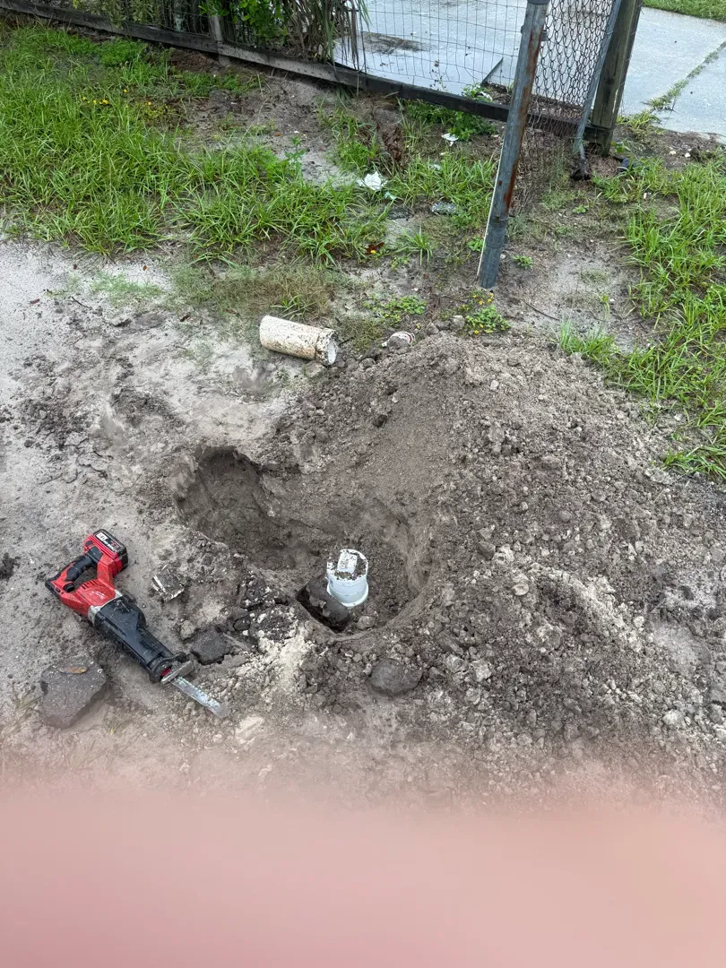 Excavated hole by a fence exposing a white PVC pipe, with a reciprocating saw on the dirt